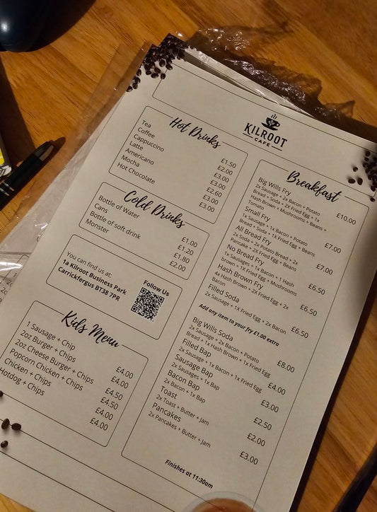 A laminated menu placed on a table, featuring various sections such as 'Hot Drinks', 'Cold Drinks', and 'Kids Menu', with pricing and description for each item.